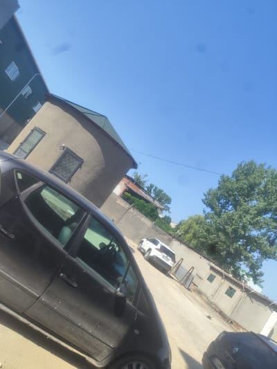 The image depicts an outdoor scene in the city of Bălți, Moldova, under a clear blue sky. The perspective is angled, looking upwards and to the right. In the foreground, a dark-colored car is partially visible, its side and windows facing the camera. To the left of the car, a two-story building with a green corrugated metal roof and several windows is visible. Adjacent to this building is another structure with a light-colored, textured facade and a dark green roof. Further into the scene, a white SUV is parked. Behind the SUV, a wall runs along the property line, with a large, leafy tree growing beyond it. The ground appears to be a dirt or gravel surface. The overall impression is that of a residential or mixed-use area.