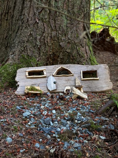 A miniature, rustic dwelling crafted from weathered wood and natural elements is positioned at the base of a large, rough-barked tree. The structure, resembling a tiny house or camper, features three openings for windows and a single door with a small, dark knob. Tiny benches and decorative elements made from twigs and moss adorn the exterior. The dwelling rests on a bed of gravel, pine needles, and small cones, with patches of moss growing on the surrounding ground and tree trunk. The scene appears to be set in a forest environment, likely a park or natural area. The location is identified as Burnaby, Canada. There are no people present in the image, and no actions are being depicted.