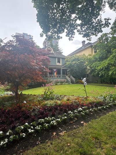 This is a photograph of a house with a front porch, set in a lush, green garden. The house is painted a muted blue-green with a cream-colored upper story. A prominent feature of the house is a pride flag displayed on the porch railing. Surrounding the house is a variety of plants, including a large, dark red-leaved tree in the foreground and a flower bed with red, white, and green foliage. The garden is meticulously maintained, with neat rows of flowers and a well-kept lawn. In the background, modern apartment buildings can be seen towering over the trees, suggesting an urban or suburban setting. The sky is overcast, indicating a cloudy or possibly rainy day. The overall mood is serene and peaceful, with a touch of vibrancy from the flowers and the pride flag. The presence of the apartment buildings hints at a location in Vancouver, Canada, where such architectural contrasts are common.