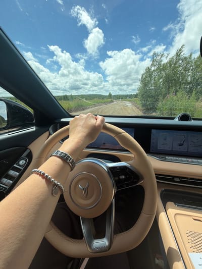 The image captures a first-person perspective from inside a car driving on a dirt road. The driver's hand, adorned with a silver bracelet and a smaller beaded bracelet with a heart charm, is on the steering wheel. The steering wheel is light tan and has the brand logo "VEYAH" at its base. The car's interior is also primarily light tan, with black accents around the door controls and dashboard.

The road ahead is unpaved and appears to be in a rural or natural setting, with green foliage and trees lining the sides. The sky is bright blue with scattered white clouds. In the distance, there are hints of more trees and a gently rolling landscape, suggesting a location outside of a dense urban area. The dashboard displays show various icons and text, indicating the car's infotainment system is active. The overall impression is a leisurely drive through a scenic, possibly remote, area.