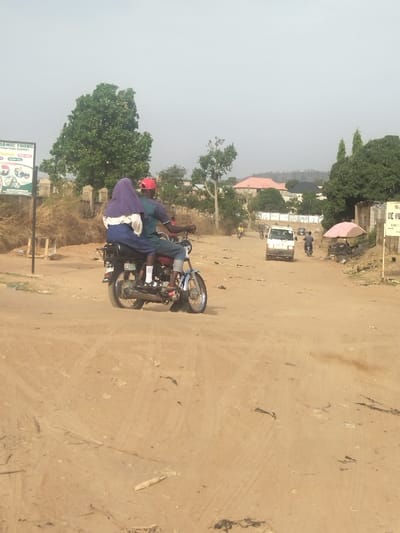 A man and a woman are riding a motorcycle on a dirt road in New Karu, Nigeria. The woman is wearing a blue hijab and the man is wearing a red hat. There is a white van in the distance and a few other vehicles are parked along the road. There are trees and bushes on both sides of the road. The ground is dusty and there are tire tracks visible. The sky is blue and there are some clouds in the distance. The road is empty except for the motorcycle. The scene is typical of a rural area in Nigeria. The image was taken in the afternoon, with the sun shining from above. The mood of the image is relaxed and peaceful.