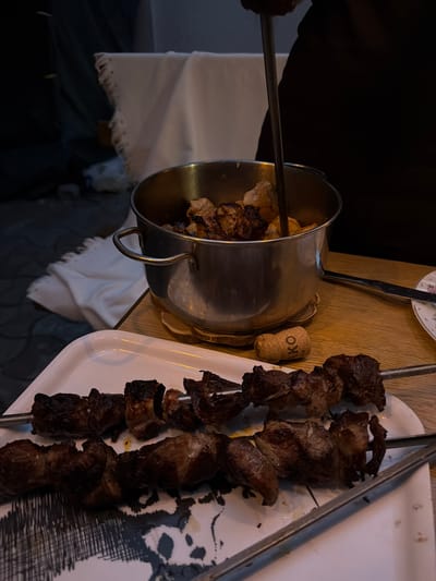 In Taganrog, Russia, a close-up shot shows a table setting with grilled meat skewers and a pot of cooked meat. The skewers, made of metal, are laden with charred pieces of meat, with two prominent skewers resting on a white platter with a decorative pattern. A metal pot, containing more cooked meat and a metal skewer extending upwards, is positioned behind the platter. The scene appears to be set on a wooden table, with a wine cork visible next to the pot. In the background, a white cloth with frayed edges is draped over a dark surface, and a tiled or paved ground is visible. The lighting suggests an indoor or evening setting. There are no visible people in the frame, but the presence of food and serving items indicates a meal in progress or recently concluded.