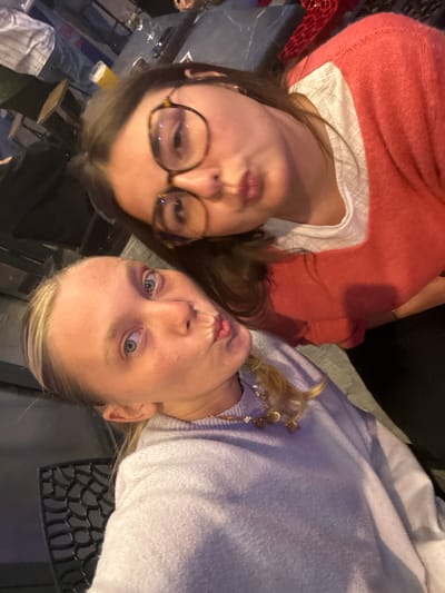 Two young women are captured in a close-up, high-angle selfie, playfully posing with pursed lips. The image is oriented sideways, with the women appearing to be lying down.

The woman on the left (wearing glasses) has medium-length dark hair and is sporting round, tortoise-shell rimmed glasses. She is dressed in a white top layered under a textured, coral-red sweater. Her lips are puckered in a "duck face" expression. Below her chin, a glass containing a amber liquid, possibly beer, is partially visible.

The woman on the right has blonde hair, which appears to be pulled back, and is wearing a soft, light purple sweater. A delicate necklace or charm bracelet with gold-colored charms is visible near her collarbone. Her blue eyes are wide open and directed towards the camera, while her lips are also pursed in a similar playful manner.

The background suggests an indoor setting, possibly a bar or casual restaurant in Lille, France. Dark surfaces, tables, and a metallic, patterned grate-like surface are visible behind them. The lighting appears artificial, indicative of an evening or nighttime setting. The overall mood is casual, friendly, and lighthearted, consistent with friends enjoying a social outing. No text is visible in the image.