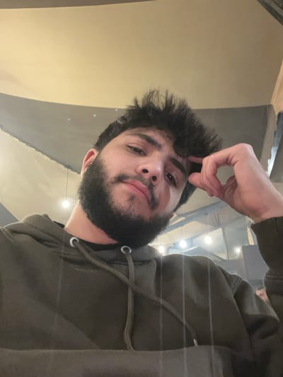 A bearded man with brown hair, wearing a green hooded sweatshirt, poses for the camera. He's holding his hand up, near his forehead, seemingly resting or scratching. The lighting appears artificial, with visible light fixtures in the background. The camera angle is slightly tilted, giving a casual, upward-looking perspective. The man's expression is calm, with a slight smirk.