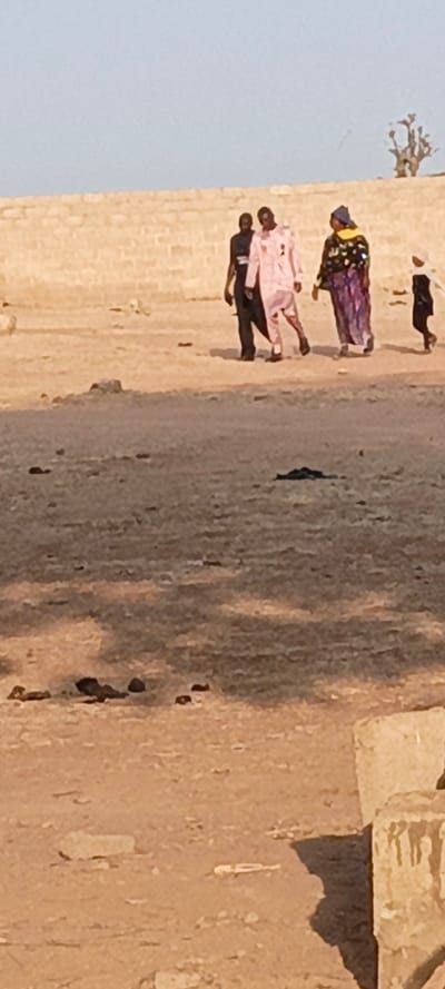 This media file captures an outdoor scene in what appears to be a dry, open environment, consistent with Rangaza, Nigeria. The image is taken during daylight hours, likely late morning or early afternoon, indicated by the clear blue sky and the distinct shadows cast on the ground.
The primary subjects are four individuals walking from left to right across the sandy, unpaved terrain. The person furthest left is dressed in a dark outfit. Next to them, a person wears a light pink or peach-colored long garment. Following them is an individual, likely a woman, adorned in a vibrant, multi-patterned dress or robe with a yellow and blue head covering. To the far right, a small child in a dark outfit walks alongside the group.
In the background, a long, light brown brick wall or structure extends horizontally across the frame. Above the right side of this wall, a sparse, dry shrub or small tree is visible against the clear sky. The ground in the foreground and mid-ground is a light brown, dusty expanse, uneven and scattered with small dark rocks and debris. Shadows, possibly from overhead branches or structures outside the frame, stretch across the ground. In the bottom right corner, a few light-colored, broken concrete blocks are visible. There is no discernible text within the image. The overall impression is of people traversing a stark, rural landscape under a clear sky.