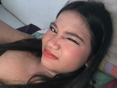 In this close-up, low-angle, portrait-oriented selfie, a young woman with long, dark hair is lying down. She is wearing makeup, including reddish-orange lipstick and blush. Her right eye is closed in a wink, while her left eye is open and looking towards the camera. Her facial expression appears playful. The background consists of a wall and bedding with a colorful striped pattern. Based on the lighting and the style of the bedding, it is plausible this image was taken indoors in a residential setting in North Jakarta, Indonesia.