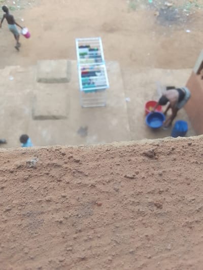 The scene depicts an outdoor ground area in Ukwu-Abwa, Nigeria, viewed from an elevated position, with a textured, granular orange-brown surface occupying the foreground. The ground is primarily exposed light brown dirt.
Three individuals are present. In the upper left, a shirtless individual in light-colored shorts walks rightward, carrying a pink plastic bucket in their left hand. To the right, another individual, wearing a dark top and light bottom, is bent over, facing left, interacting with three plastic basins on the ground: a red one, a blue one, and a lighter blue/white one. Further to the lower left, a third, heavily blurred individual wearing a blue top is partially visible.
Centrally located is a white metal clothes drying rack, a multi-tiered structure, with various multicolored garments hanging on its rungs. Two rectangular concrete slabs are embedded in the dirt ground to the left of the drying rack. A section of an orange-brown building wall is visible on the far right. The background shows more dirt ground with minimal, indistinct vegetation or debris.