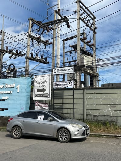 In Kathu, Thailand, a silver Ford Focus sedan is parked on a paved road next to a light blue building with the words "THE VALLEY" and the number "1" visible. A large utility pole with numerous electrical wires and transformers dominates the background. Several signs are attached to the utility pole. One sign, in black and white, reads "ห้องพัก รายเดือน" (rental rooms monthly) with an arrow pointing to the right and a phone number "081-370-3355". Another sign, with a cat graphic, says "Fur seasons" and below it "cat grooming & hotel". A third sign, partially obscured, displays the text "Babushka" and "home made with care". A concrete wall with graffiti runs behind the car and the utility pole. The sky above is partly cloudy. Two white notices are affixed to the driver's side windows of the car.