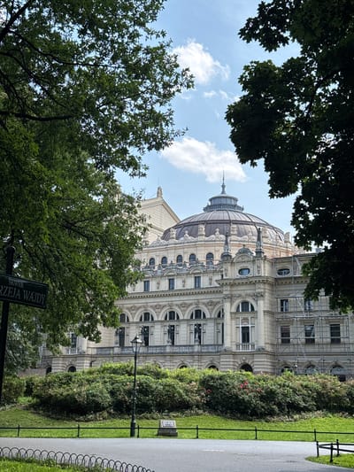 The image shows a partial view of the Juliusz Słowacki Theatre in Krakow, Poland, on a partially cloudy day. The theatre is a grand building with a domed roof, ornate facade, and classical architectural elements. It is surrounded by lush green trees and manicured bushes. In the foreground, a street sign with Polish text and a black metal fence can be seen. The scene suggests a calm, pleasant day in a park or public square, with the theatre as the prominent focal point.
