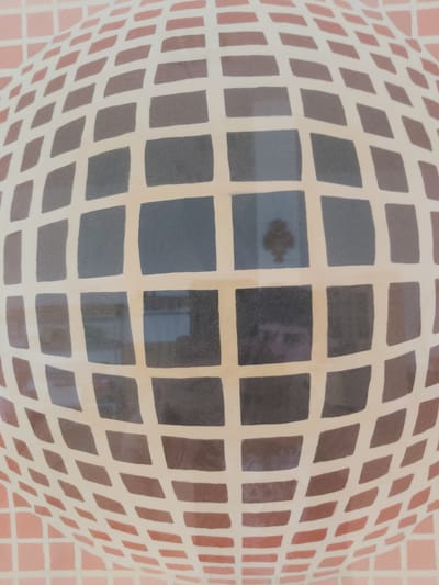 The image depicts a close-up, distorted view of a tiled surface, likely a wall or floor, with a grid pattern. The tiles are squares of varying shades of light brown, dark gray, and pink. The grid lines are a uniform light cream color. The distortion suggests the surface is curved, possibly a dome or sphere, or the image was captured with a wide-angle lens. Reflections are visible within some of the darker tiles, hinting at a surrounding environment, possibly indoors. In one of the dark tiles, a faint reflection of a person's face and hair can be seen. Another dark tile shows a reflection of a dark, four-pointed object against a light background. The image does not contain any recognizable landmarks or features that would allow for precise identification of its location within the city of Tryavna, Bulgaria.