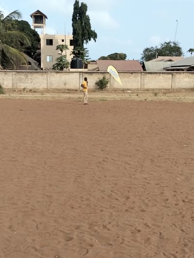 A man stands on a large, sandy field near a yellow advertising flag. Behind him is a low brick wall with graffiti and various buildings, including one with a tower and another with a water tank. The scene is bathed in daylight under a clear, blue sky. The ground is a light brown color, covered in sand and appearing dry.