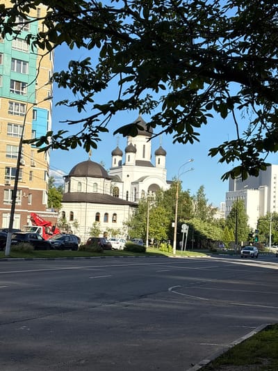 In Mytishchi, Russia, a sunlit street scene unfolds with a prominent church featuring a dark dome and white facade, partially obscured by the dark green leaves of a tree in the foreground. To the left, a tall residential building with turquoise and yellow brickwork stands, while to the right, a modern white building and a cluster of trees line the road. Cars are parked along the side of the street, and a red crane truck is visible. Pedestrians can be seen walking on the sidewalk in the distance, and traffic lights are visible at an intersection. The sky is a clear blue.