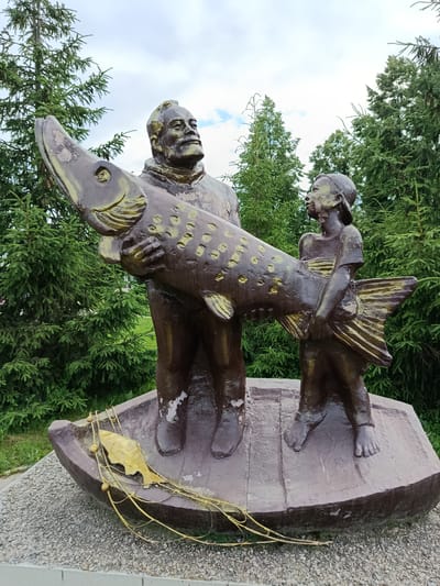 This is a full outdoor shot of a bronze sculpture in Elovo, Russia, depicting two figures, an adult and a child, standing in a boat and holding a large fish. The sculpture is situated on a gravel base surrounded by evergreen trees. The adult, a man with a mustache, is positioned behind and to the left of the child. He is holding the head of the fish, which is positioned horizontally. The child, wearing a baseball cap, is to the right of the adult and holds the tail of the fish. Both figures are looking upwards and to the right. In the boat with them is a fishing net and a smaller golden fish. The sky is overcast with some clouds. The sculpture has a weathered appearance with areas where the bronze is chipped.