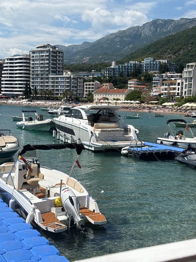 The image depicts a scenic view of a coastline in Rafailovići, Montenegro, characterized by a picturesque beach lined with numerous sun umbrellas and beach chairs, with a scattering of people enjoying the seaside. Behind the beach, modern, multi-story buildings and some older structures form a dense urban landscape that stretches along the coast. The buildings vary in architectural style, from sleek, contemporary designs with large windows to more traditional facades with red-tiled roofs.
In the foreground and middle ground, several boats are moored in clear, blue-green water. These include a large white motor yacht with the text "LEADER 40" visible on its hull, and other smaller vessels, some of which appear to be speedboats or tenders. A dark blue floating dock is visible to the right of the frame, connected to the boats. The water is calm and transparent, allowing a view of the seabed in some areas.
Towering over the coastal town and the water is a large, lush green mountain. The mountain's slopes are densely covered with trees, and its rocky terrain is visible in places. The sky is partly cloudy with scattered white clouds against a bright blue backdrop, suggesting pleasant weather, likely during the daytime, possibly mid-morning or early afternoon. The overall impression is one of a vibrant tourist destination, combining natural beauty with urban development and recreational activities on the water.