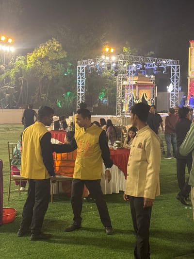 A nighttime event displays a stage structure with lighting and decor. Several people are present, including three men prominently dressed in yellow and black outfits. One man is wearing a beige jacket. They are standing on green artificial turf. Tables with red tablecloths and various event items are visible in the background. Trees and additional structures with lighting are also present.