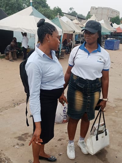 Two adult females are standing and conversing outdoors in Ura Nta Umuarandu, Nigeria. The ground is an uneven, light brown dirt surface.
The female on the left wears a light blue long-sleeved collared shirt, a black pencil skirt, and black flat shoes. Her hair is braided and styled into a bun. She wears a small hoop earring in her visible ear and carries a black backpack on her left shoulder. She holds a white plastic bag in her right hand and points her left hand downwards.
The female on the right wears a black baseball cap, a white polo shirt with blue trim and an "AbiaFm" logo on the left chest, a faded blue and yellow-brown denim midi skirt, white athletic sneakers, and white socks with blue stripes. She wears a light-colored wrist watch on her left wrist. She holds a white tote bag containing papers in her left hand and a dark-colored smartphone in her right hand.
Both women are facing each other, engaged in interaction.
In the background, numerous pointed tents, primarily white with blue or green stripes, are erected, suggesting an outdoor market or event. Under a tent to the far left, several men are seated. To the right, a multi-story concrete building with multiple windows is visible. A blue rectangular container is also present in the mid-ground. The sky is overcast.