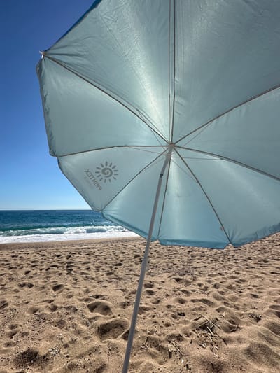 This image depicts a tranquil beach scene in Pineda de Mar, Spain, observed from a low-angle perspective beneath a beach umbrella. A large, light blue beach umbrella, illuminated from above, occupies the foreground and mid-ground. Its translucent fabric reveals the internal framework of white support spokes and a central pole. Reversed text "PRINTEX SUMMER" along with a sun logo is discernible on the underside of the canopy. The umbrella is anchored in light brown, coarse sand, which forms the lower half of the image and displays numerous depressions and subtle undulations. Beyond the sandy expanse, the Mediterranean Sea exhibits moderate wave activity, with white foam visible at the shoreline. The sea transitions from turquoise near the beach to a deeper blue towards the horizon. A clear, unblemished blue sky occupies the upper left quadrant of the frame. No individuals, animals, or specific actions are visible within the scene.