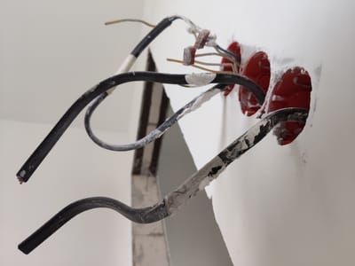 The image shows a close-up view of electrical wiring in a building under construction or renovation. Several black electrical cables, some with exposed copper wire ends, are visible. They are connected to junction boxes embedded in a white wall, which appear to be painted red. The wall has a rough texture, suggesting it may be plaster or drywall that has not yet been fully finished. Some of the cables are wrapped with white tape, and there are remnants of white putty or plaster around the junction boxes. The scene is indoors, with artificial lighting. The overall impression is one of unfinished electrical work. No people or text are visible in the image. The location context is Ходаево, Russia.