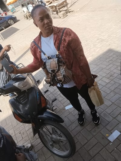 An outdoor scene in Fagge A, Nigeria, features an adult female standing on a light-colored paved surface, looking towards the left of the frame. She wears a white t-shirt with text, a reddish-brown long-sleeved shirt over it, black pants, and black sneakers with white soles and stripes. A plaid backpack (beige, brown, black pattern) with dark straps is worn on her back. She holds a light brown paper bag in her right hand and a dark smartphone in her left hand, with an earbud visible in her left ear. A dark motorcycle, partially covered with a light fabric on its handlebars, is parked to her left. Another individual is partially visible to the bottom left, next to the motorcycle, holding a smartphone. In the background, a street is visible with a blue car parked, alongside several wooden market stalls and two distant figures seated. The area is illuminated by strong overhead sunlight, casting shadows.