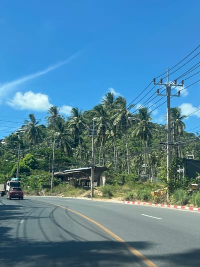 The image shows a road in Baan Chaweng Noi, Thailand, with a lush, tree-covered hillside in the background. The sky is a vibrant blue with a few wispy clouds. Coconut palm trees dominate the vegetation on the hillside, with some buildings and power lines visible among them.
In the foreground, the asphalt road has yellow and white lane markings. Tire tracks are visible on the road surface. A red car is driving on the road, followed by a white truck. The roadside is lined with red and white painted curbs and dense green vegetation. A small, rustic wooden structure with a corrugated roof is situated near the roadside. The overall impression is a tropical, somewhat undeveloped area, likely a coastal region given the typical scenery associated with Thailand.