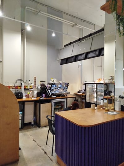 This image depicts the interior of a cafe or restaurant. In the foreground, a curved wooden counter is prominent, with a blue corrugated base. A black chair with a metal frame is positioned in front of the counter. Behind the counter, a glass display case showcases pastries or food items. Various coffee-making equipment, including an espresso machine and grinders, are visible. Bottles of beverages are arranged on a shelf behind the counter. The walls are painted in light colors, and overhead lighting fixtures are present. A ventilation system is installed along the ceiling. A person with red hair is partially visible, seated or leaning over the counter. The overall environment suggests a commercial food and beverage establishment, likely in an urban setting. The specific location in the city of Kommunarka, Russia, is not discernible from visual cues within the image itself.