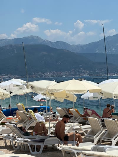 A busy beach scene unfolds in Baošići, Montenegro, under a clear blue sky dotted with fluffy white clouds. Numerous white and cream-colored beach umbrellas provide shade for lounge chairs occupied by people relaxing on the sand. The water is calm and appears to be a vibrant turquoise. In the background, sailboats with tall masts are moored, and further out, a town with buildings lines the coast. Beyond the town, a range of imposing, blue-toned mountains forms the horizon.
In the foreground, several individuals are visible. A woman in pink bikini bottoms and a black top sits on a lounge chair, holding a drink and a phone. Beside her, a man with dark hair and shorts is seated, looking away. Further back, another man reclines on a lounge chair. A woman in a bikini is seen in the water, floating on a pink inflatable. Other beachgoers are scattered across the scene, some lying down, others conversing. The overall impression is one of a lively and pleasant summer day at the beach.