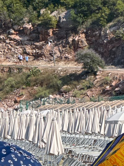 The image depicts a coastal scene in Kamenovo, Montenegro, characterized by a rocky hillside and a beach lined with numerous closed beach umbrellas. In the background, a steep, rocky incline covered with sparse vegetation and trees dominates the landscape. A paved pathway runs horizontally across the mid-ground, upon which two individuals are walking. One person, wearing a light purple top and dark shorts, is standing near a park bench. Beside them, a person in a blue t-shirt and dark shorts is walking away. The pathway is bordered by a low stone wall and further down, by dense green scrub. The foreground is occupied by a beach with rows of closely packed, light-colored beach umbrellas, mostly closed and standing upright. A few beach chairs are visible between the umbrellas. In the lower left corner, a partially visible open blue umbrella with white anchor patterns is present, and to the right, a portion of a colorful umbrella or shade structure is visible. The overall impression is a sunny day at a beach resort, with the natural rocky terrain of Montenegro forming a dramatic backdrop.