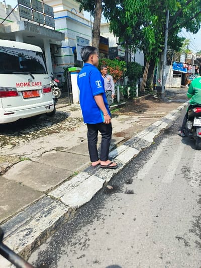 The image shows a man in a blue shirt with black pants standing on a sidewalk in Cimahi, Indonesia.  He is standing near a white van with the words "HOME CARE" printed on the side. The van is parked on the side of the street, facing the camera. The man is looking to the right and appears to be talking to another man. The second man, who is wearing a gray shirt, is standing behind the man in blue and facing the opposite direction.  The sidewalk has a concrete curb that separates it from the street. The street is asphalt and has several faded white lines painted on it. There is a motorcycle in the background, partially visible in the right side of the image.  The motorcycle is driven by a man who is wearing a green shirt. The motorcycle is facing left and is about to cross the intersection. The image appears to be taken on a sunny day.  The area is residential.  The buildings in the background are painted white and have green trim.  There is a tree in the foreground.
