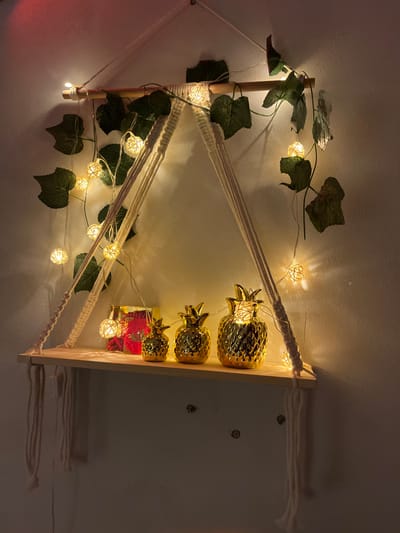 This image depicts a decorative macrame hanging shelf display, situated indoors in Nuseirat, Palestinian Territory. The central feature is a light-colored wooden plank shelf, suspended by cream-colored macrame cords that form two triangular supports from a horizontal wooden dowel. The dowel is attached to a single top cord, fastened to an unseen point above.

On the shelf, three golden, textured pineapple-shaped figurines are arranged in decreasing size from right to left. To the left of the smallest pineapple, a small, rectangular red object with yellow details, possibly text or a design, is partially visible.

Above and around the shelf, a string of warm white LED fairy lights, encased in spherical, woven rattan-like covers, is draped. These lights are interspersed with artificial green ivy leaves that twine around the wooden dowel and hang down, creating a lush, illuminated effect. The entire arrangement is set against a plain, light-colored wall, which appears to be off-white. The scene is dimly lit, with the primary illumination originating from the fairy lights, casting soft glows and shadows. Two small, dark, circular objects, possibly screw heads or anchors, are visible on the wall below and to the left of the shelf.