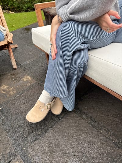 The image displays a close-up, downward-angled view of a seated individual's lower body in an outdoor setting in Huasca de Ocampo, Mexico. The person wears a grey knit sweater with visible white stripes on the sleeves, blue and white vertically striped wide-leg trousers, cream-colored ribbed socks, and tan suede clogs with a brass-toned buckle on the strap. A silver bracelet adorns the left wrist, and multiple rings are present on the fingers, including a silver band on the right ring finger and a ring with a turquoise-like stone on the left index finger. The individual's hands are resting in their lap, with fingers interlocked or loosely placed. They are seated on a modern wooden bench or sofa featuring a cream-colored upholstered cushion. To the left, a portion of another light-colored wooden slatted outdoor chair is visible. The ground beneath the subject is composed of dark grey, textured paving stones or flagstones, which appear damp or wet, reflecting some light. In the blurred background, vibrant green grass and dense foliage are present, suggesting a garden or patio environment.