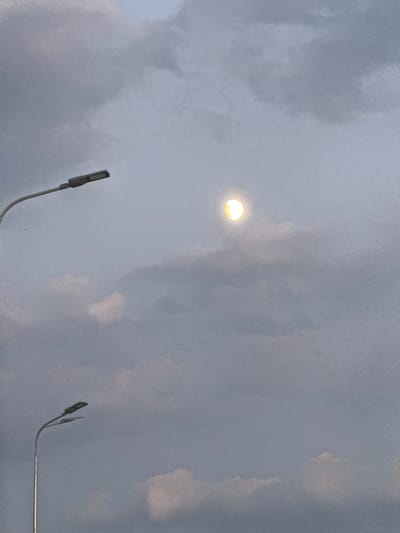 The image displays a cloudy sky with two streetlights visible. One streetlight is positioned in the upper left quadrant, and the other is in the lower left quadrant, appearing slightly closer to the viewer. In the upper central portion of the sky, a luminous, round object, likely the moon partially obscured by clouds, emits a soft yellow glow. The clouds are dense and appear to be a mix of darker and lighter gray tones, suggesting overcast conditions or approaching dusk. There are no people or discernible actions captured in the scene. The overall environment appears to be an outdoor urban setting. The location of Berezhany, Ukraine, is not visually discernible from the provided image itself.