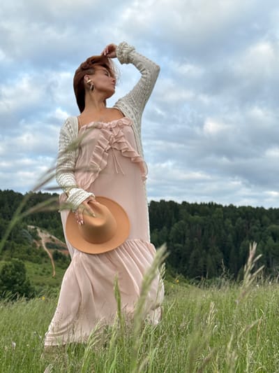 A woman with auburn hair is standing in a field of tall green grass, wearing a light pink ruffled dress and a cream-colored knitted cardigan. She is holding a tan fedora hat in her right hand, which is resting on her hip. Her left arm is raised, with her hand touching her hair. Her head is tilted back, and she is looking upwards. Behind her, a dense forest of dark green trees stretches across a rolling hillside, with a pale, overcast sky above. The location is identified as Olkhovka, Russia, suggesting a rural or semi-rural setting.