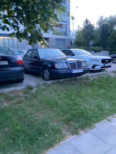 The image shows a street scene in Warsaw, Poland, with several cars parked along a sidewalk. In the foreground, a dark-colored sedan is parked next to a light-colored SUV. Both vehicles are facing away from the camera. Behind the parked cars, a building with white brick facade and large windows is visible. Trees with green foliage are present in the background. The weather appears to be overcast. The license plate of the dark sedan reads "WP 887 GP". The license plate of the white SUV is partially obscured but appears to start with "WGD".