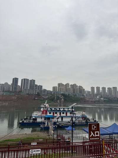 The image shows a waterfront scene in Shapingba District, Chongqing, China. Several passenger boats are docked, with a visible "Entrance" sign in English and Chinese characters. In the background, numerous high-rise apartment buildings line the riverbank. The sky is overcast. A small aircraft is visible in the distance.  A red metal fence runs along the foreground.  Signage indicates "Queue in Order".