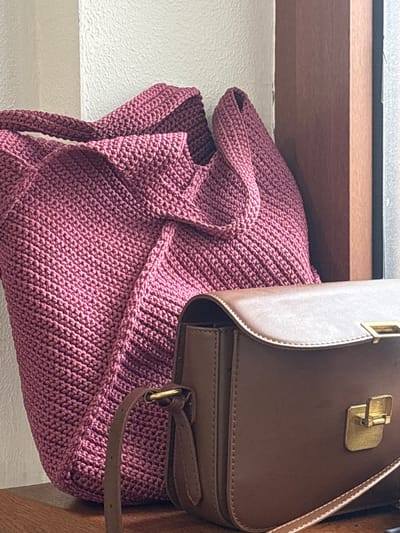 In an indoor setting, two bags are placed on a wooden surface. On the left is a large, magenta, crocheted tote bag. Its texture is dense and exhibits a diagonal stitch pattern. To the right of the tote is a smaller, brown, faux-leather crossbody bag with a flap closure and gold-toned hardware. The background consists of a textured off-white wall on the left and a dark brown wooden frame on the right, suggesting a window or doorway. The lighting is soft, creating gentle shadows. The location is Estosadok, Russia.