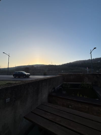 The image depicts an early morning scene in Grevenmacher, Luxembourg, with the sun rising over distant hills. The sky displays a gradient from a warm yellow-orange near the horizon to a clear light blue overhead.
In the midground, a dark grey SUV, likely an Audi Q2, travels left to right on a multi-lane asphalt road. The road surface appears to have a slight sheen, possibly from frost or dampness. Several streetlights with dark poles and curved arms are positioned along the road and ascend the hillside in the background. A small red and white triangular sign is visible on the right side of the road, adjacent to a streetlight. The background consists of rolling hills covered with sparse, possibly frosted, trees and vegetation, silhouetted against the rising sun.
The foreground is dominated by a weathered concrete structure, functioning as a retaining wall or barrier, running horizontally across the lower third of the frame. Behind this concrete structure, to the right, a rectangular water feature or pond contains dark water and green aquatic plants, including what appear to be reeds. A dark pipe or hose is visible on the left edge of this water feature. In the lower right corner, a dark, slatted wooden surface, consistent with a bench or table, is partially visible, cast in shadow.