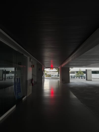The image shows a dimly lit hallway in a building. The hallway is long and narrow, with a dark wall on the left and a lighter wall on the right. A window is visible at the end of the hallway, providing a view of trees and another building. There are some objects on a shelf below the window, including a first aid kit and a box. There is a digital display showing numbers in red at the end of the hallway. This scene appears to be within the city of Heroica Puebla de Zaragoza, Mexico, as indicated by the location context.