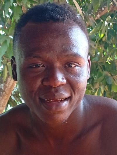 A close-up portrait features a smiling Black man with short, dark hair. He is outdoors, with green foliage visible in the background. His skin appears smooth and reflects light. His teeth are slightly visible. The focus is sharp on his face, emphasizing his expression. The lighting is natural and bright.