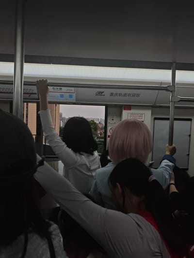 The image shows the interior of a Chongqing Rail Transit train in China. A sign above the window identifies the station as "Shijingpo" in the Shapingba District. Several people are visible inside the train, including three women in the foreground. One woman, wearing a white long-sleeved shirt, has her arm raised, holding onto a pole. A second woman has pink hair, and a third woman has her hair tied in a ponytail. The image is taken from the perspective of another passenger on the train. Outside the window, city buildings and trees can be seen.