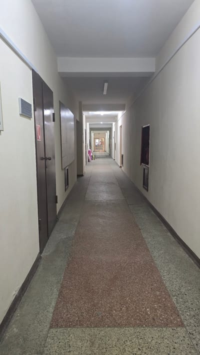 This media file shows a long, brightly lit corridor looking down from a high angle, giving a sense of extreme perspective. The corridor appears to be in a building with multiple floors, as suggested by the receding openings in the ceiling and the repeating pattern of doors and walls. The walls are a neutral color, and the floor is covered in a speckled terrazzo material, with a large rectangular inlay of reddish-brown terrazzo in the center.
The corridor is sparsely furnished. In the foreground, there are several dark brown doors, one of which has a red and white fire exit sign visible. There are also some flat, dark surfaces that could be tables or shelves. Further down the corridor, near the center, are a few items that appear to be stacked luggage or bags, in shades of pink and white. At the far end of the corridor, a set of glass doors leads to another section of the building, which is also illuminated.
The overall impression is one of institutional architecture, possibly a dormitory, apartment building, or hospital. The lighting is bright and even, suggesting it is daytime. There is no visible weather, as the scene is entirely indoors. The extreme perspective, with the corridor narrowing to a single point in the distance, creates a dramatic visual effect. The scene is devoid of people, contributing to a sense of stillness and perhaps slight emptiness.
Some text is visible: a red and white fire exit sign on a door in the foreground. Another small, rectangular sign with white text on a blue background is visible on a white surface in the foreground.
