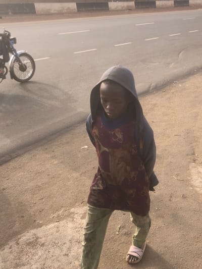 A young person stands on an unpaved dirt and gravel shoulder adjacent to a paved road in Hanwa, Nigeria. The individual wears a grey hooded sweatshirt with the hood up, a maroon long-sleeved shirt patterned with a light-colored animal print, light-colored trousers, and a single light pink slide sandal on their right foot. Their head is tilted downwards, obscuring their facial expression. In the left background, a light-colored motorcycle is partially visible on the asphalt road, casting a shadow. The paved road features faded white dashed lane markings. A low, dark structure with a black and white striped top is visible in the distant background. The scene is illuminated by natural daylight, and the atmosphere appears slightly hazy.