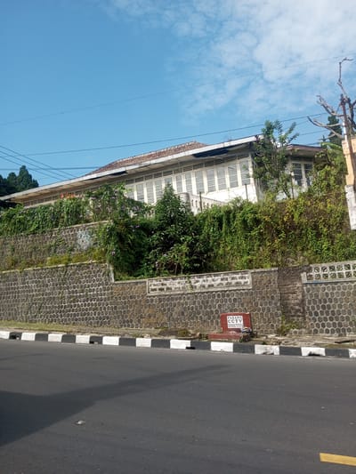 The image shows a two-story white building situated on an elevated stone foundation. Lush greenery covers parts of the wall and the area around the building. The architecture suggests an older style with prominent windows. In front of the wall is a paved road with a black and white curb. A small red rectangular sign with the text "PASANG CCTV" and a phone number is visible in front of the wall. The sky is a clear blue with scattered clouds. The scene appears to be in a residential area on a sunny day. The location is implied to be Khe Luang, Laos.