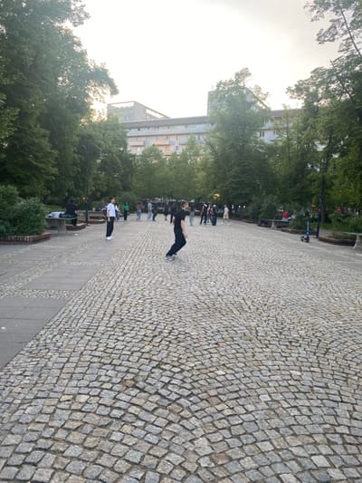 The image shows a cobblestone courtyard located in the city of Warsaw, Poland, at the coordinates 52.234 latitude and 21.007 longitude. There are several people standing and walking around the courtyard. A single person is seen kicking a ball, which is in mid-air, towards the left side of the image. The courtyard is surrounded by trees and apartment buildings. In the background, a light blue electric scooter is parked on the cobblestones. There are also some benches in the background to the right.  The sky is overcast with gray clouds. The image is taken from a low angle. The cobblestones are gray and brown, and the courtyard is empty except for the people.
