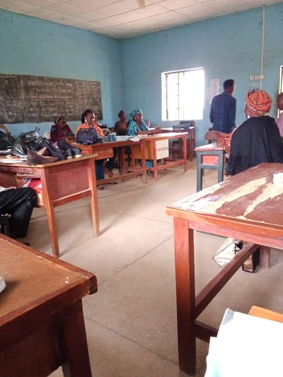 The image depicts an indoor scene, likely a classroom or meeting room, in Kakuri Gwari, Nigeria. Several people, predominantly women, are seated at wooden desks. One woman in the foreground on the right wears a black garment and an orange headwrap, facing away from the camera. Another woman, also in the foreground on the left, wears a black outfit and a colorful headwrap. Other individuals are visible seated at desks further back in the room.

The room has light blue walls and a tiled floor. A blackboard with handwritten text is visible on the far wall, along with a window letting in natural light. There are several wooden desks and chairs, some of which appear to be older and well-used, with visible wear and tear. Some desks have bags and other items placed on them.

The blackboard contains handwritten text, including what appears to be contact information, account details (FCMB 1031409295), and phrases like "SCHOOL WELFARE REMITTANCE" and "EVIDENCE & PAYMENT". The mention of "AGILE SPIN 2024" is also visible.

The overall impression is of a gathering or meeting taking place in a functional but not modern setting. The lighting suggests it is daytime. The expressions of the people are not clearly discernible, but the atmosphere appears calm and focused on the activity at hand.