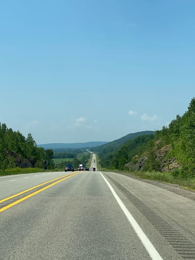 The image shows a highway scene in Pointe-au-Chêne, Canada, on a clear, sunny day. The camera is positioned from a vehicle traveling on the highway, looking forward. The road is paved asphalt with yellow double lines marking the center and a white line along the right edge. The highway is a two-lane road, with traffic visible in the distance.
On the left side of the road, there is a steep, rocky embankment covered in dense green trees. On the right side, the terrain slopes upwards, also covered in lush green trees and rocky outcrops. The landscape in the distance consists of rolling hills and fields, suggesting a rural or semi-rural setting. The sky is a clear, pale blue with a few scattered white clouds.
Several vehicles are on the highway in the distance. Closest to the camera, on the right lane, is a dark-colored car. Further ahead, a few other cars and what appears to be a white truck are visible, all traveling in the same direction. The perspective of the image emphasizes the long, straight stretch of the highway disappearing into the horizon, framed by the natural landscape. The overall impression is one of a scenic drive on a clear day in a wooded, hilly region.