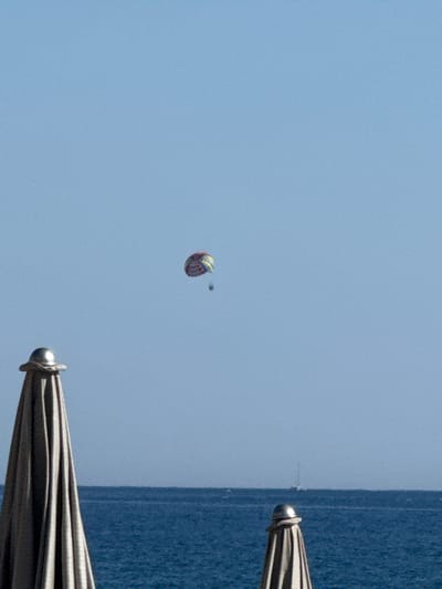 A parasailer is in the sky above the Adriatic Sea off the coast of Rafailovići, Montenegro. The parasailer is visible as a small, colorful canopy with two people beneath it, suspended in the blue sky. In the foreground, two beach umbrellas, folded and covered, are positioned to the left and right of the frame. The sea is a deep blue, and a small sailboat is visible on the horizon. The sky is clear and bright blue.