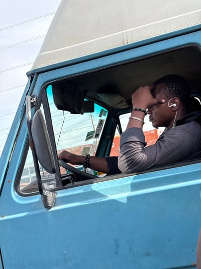 An adult male is observed in the driver's seat of a light blue commercial vehicle, possibly a van, in Igboefon, Nigeria. The individual, with dark skin, wears a dark long-sleeved shirt. A white wired earbud is present in his right ear, and his left wrist is adorned with a dark beaded bracelet and a silver-colored bracelet. His left hand rests on the dark steering wheel, while his right hand is pressed against his forehead, obscuring the upper facial features as he gazes towards the right side of the vehicle. The vehicle exhibits a light blue exterior with visible wear, including rust near the top left of the windshield frame, and an elevated, off-white roof. A large, dark side mirror is affixed to the driver's door. The interior headliner is dark, and multiple decals are visible on the inside of the windshield. The background, viewed through the vehicle's windows, shows a bright sky with overhead wires and indistinct structures.