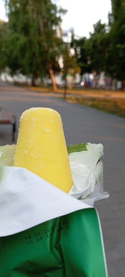 A close-up shot features a bright yellow popsicle partially unwrapped from its white and green packaging. The popsicle is the main subject, with its textured, frosted surface catching the light. The packaging, made of a reflective material, is visible in the foreground, with some of its layers peeled back. In the blurred background, a pathway or road is visible, lined with trees. The golden light suggests it's either early morning or late afternoon. The overall impression is a casual moment, possibly outdoors, enjoying a frozen treat.

Visible text on the packaging includes "16.02.2023 03:52" and "10.08.2023 VB 2". This text likely indicates a production or expiry date and a batch number. The setting appears to be a park or an outdoor public space, with trees and a path suggesting a natural or recreational environment. The soft focus on the background elements draws attention to the popsicle itself. The location context is Kirsanov, Russia, which could imply the type of trees and general flora visible in the background.