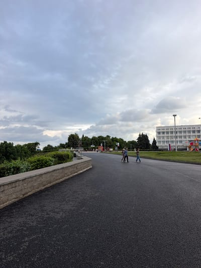 The image shows an outdoor scene with a paved area in the foreground and a building in the background. Several people are walking on the paved area. There are trees and greenery along the side of the paved area, separating it from the building. The building appears to be an institutional or office building, with multiple windows. There is a playground structure visible near the building. A flag, possibly the Serbian flag given the location context, is visible on the building. The sky is cloudy, suggesting a potentially overcast day. The time of day appears to be daytime. The scene seems to depict a public space or an area adjacent to a large building.
