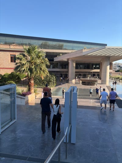 The image shows a group of people walking along a paved walkway in front of a modern building with large glass windows. The building's architecture is contemporary, with clean lines and geometric shapes. Some people are dressed casually, suggesting a warm day, possibly afternoon, given the bright sunlight casting shadows. The background reveals a clear blue sky, indicating good weather.
The scene appears to be a public space, possibly a museum or a cultural center, given the architectural style and the presence of visitors. Some individuals are walking towards the camera, while others are in the distance, interacting with the environment. A palm tree is visible in the mid-ground, adding a touch of greenery and suggesting a Mediterranean climate, consistent with the provided location context of Athens, Greece.
Notable details include the varied attire of the people, ranging from t-shirts and shorts to more formal wear. The ground is covered in large, grey tiles with a marble-like pattern. A metal railing is visible on the right side of the image, and a section of a metal detector frame is present in the foreground, hinting at security measures in place. There is no visible text in the image. The overall mood seems relaxed and leisurely, with people enjoying their time outdoors.